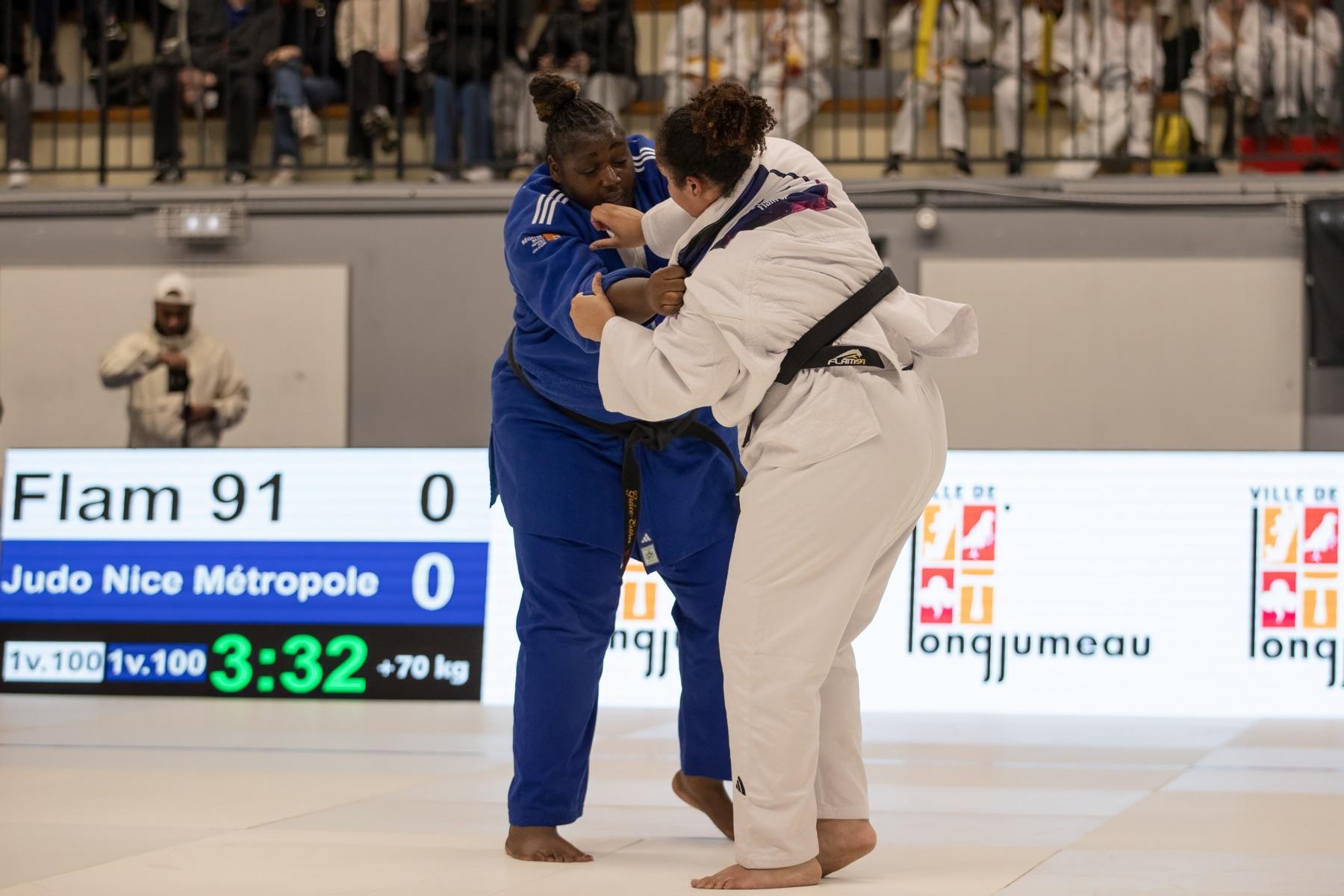 Une judokate de Judo Nice Métropole en judogi bleu au contact d'une adversaire du FLAM 91, devant le tableau d'affichage de la rencontre à Longjumeau